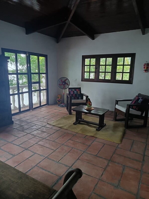 A room with a tiled floor, a fan, a table with a decorative item, a sofa, and a window with a view of greenery.