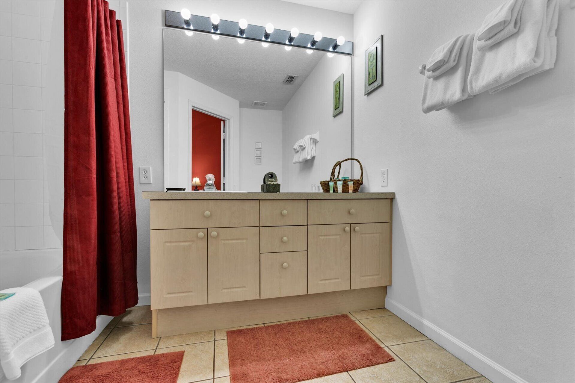 A bathroom with a vanity, a mirror with lights, a red shower curtain, and a white towel on the wall.