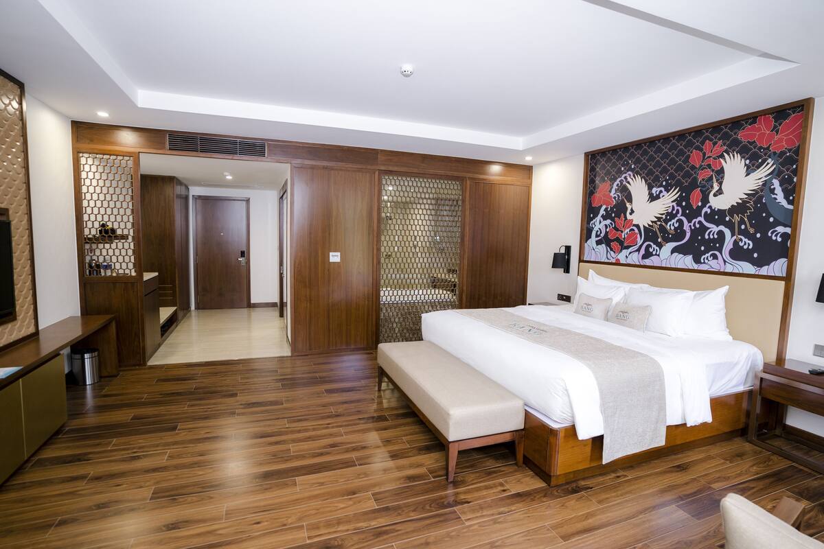 A modern hotel room with a wooden floor, a large bed with a bench, a TV, and a decorative wall painting.