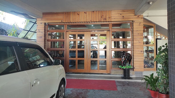 A building entrance with a white car parked outside, wooden paneling, and potted plants. - Hotel Sanjivani Heritage (Dibrugarh)