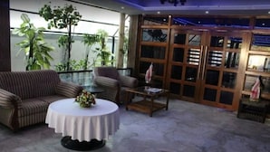 A lounge area with striped sofas, a round table with a floral centerpiece, and a view of indoor plants through large windows. - Hotel Sanjivani Heritage (Dibrugarh)