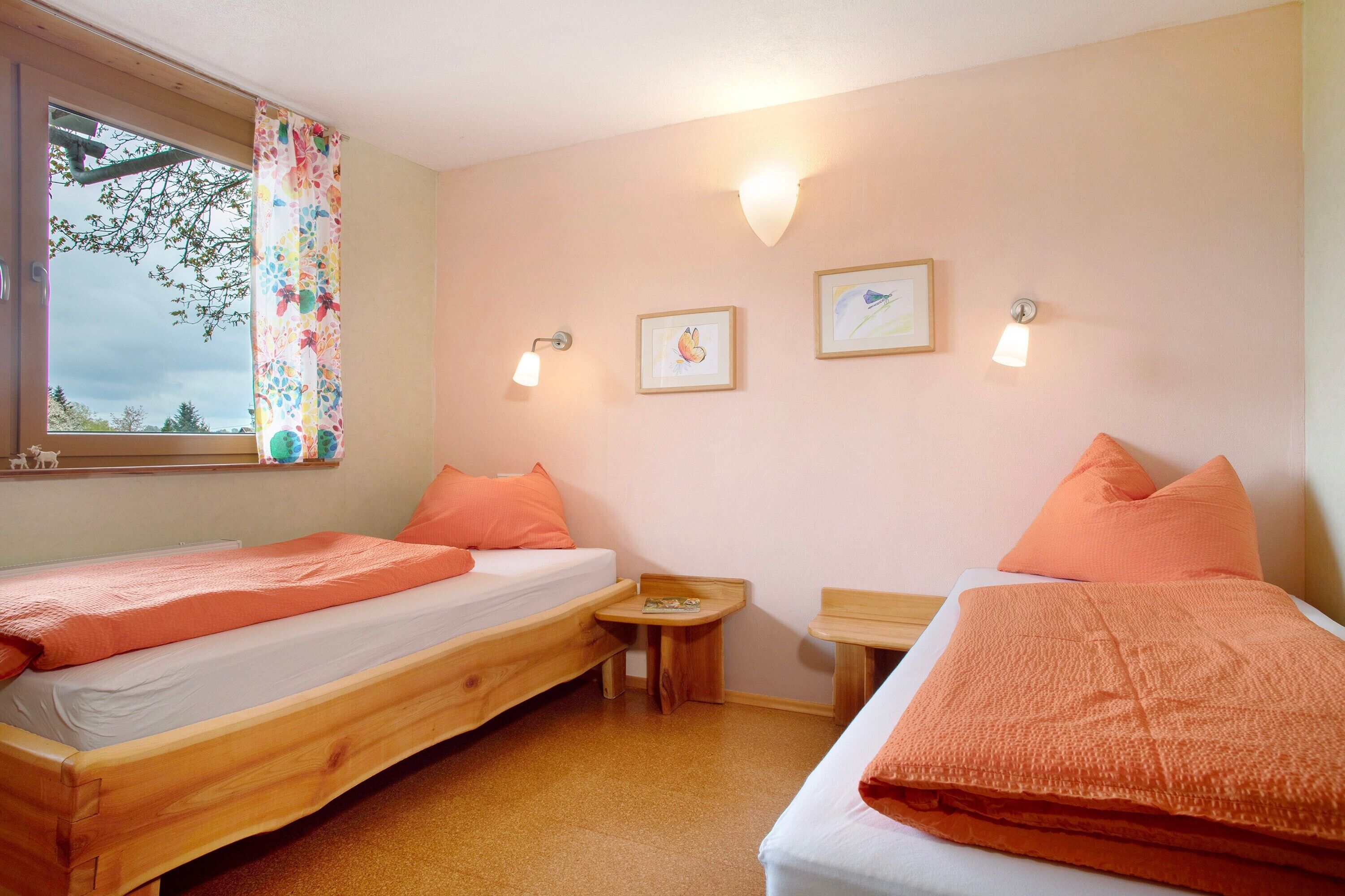 A room with two wooden beds, a small table, a window with a curtain, and wall-mounted lights.