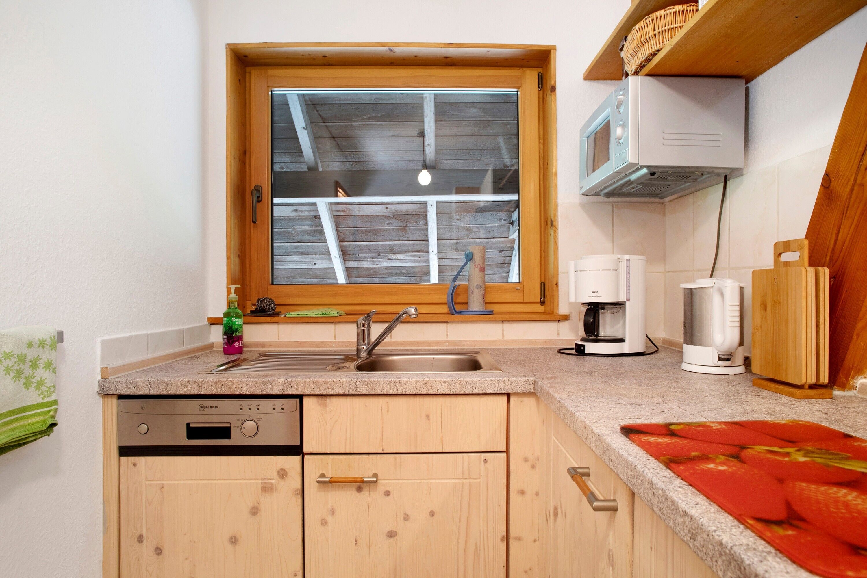 Eine Küche mit Fenster, Holzschränken, einem Geschirrspüler, einer Kaffeemaschine und einer Mikrowelle.