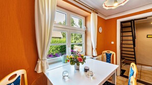 A room with a white table, chairs, a window with curtains, a vase with flowers, and a view of greenery outside. - Holiday Apartment 'Fachwerkferienhaus' with Lake View, Terrace & Wi-Fi (Ulrichshusen)
