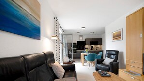 A modern living room with a black leather sofa, a wooden coffee table, and a dining area with a blue chair. - Holiday Apartment 'Im Naturpark Rhein-Westerwald' with Private Terrace and Wi-Fi (Puderbach)
