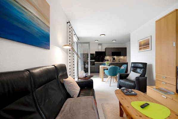 A modern living room with a black leather sofa, a wooden coffee table, and a dining area with a blue chair.