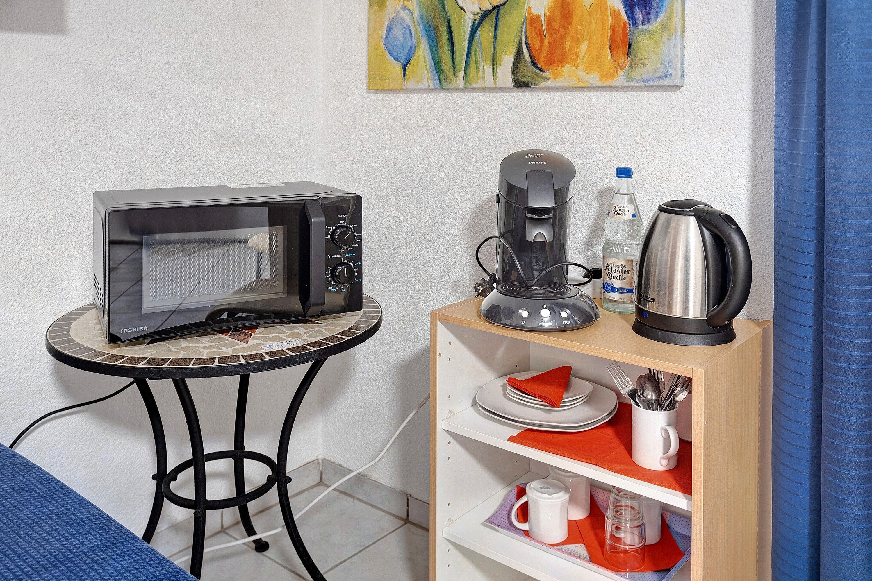Une petite cuisine équipée d’un four à micro-ondes, d’une machine à café, d’une bouilloire et d’une étagère contenant des assiettes, des tasses et des ustensiles.