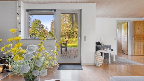 A modern interior with a wooden floor, a dining area with a table and chairs, a sofa, and a view of a garden through a sliding door.