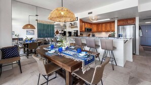 Une cuisine moderne avec des meubles en bois, un îlot central et un coin repas agrémenté de chaises en osier et d’une nappe bleue.