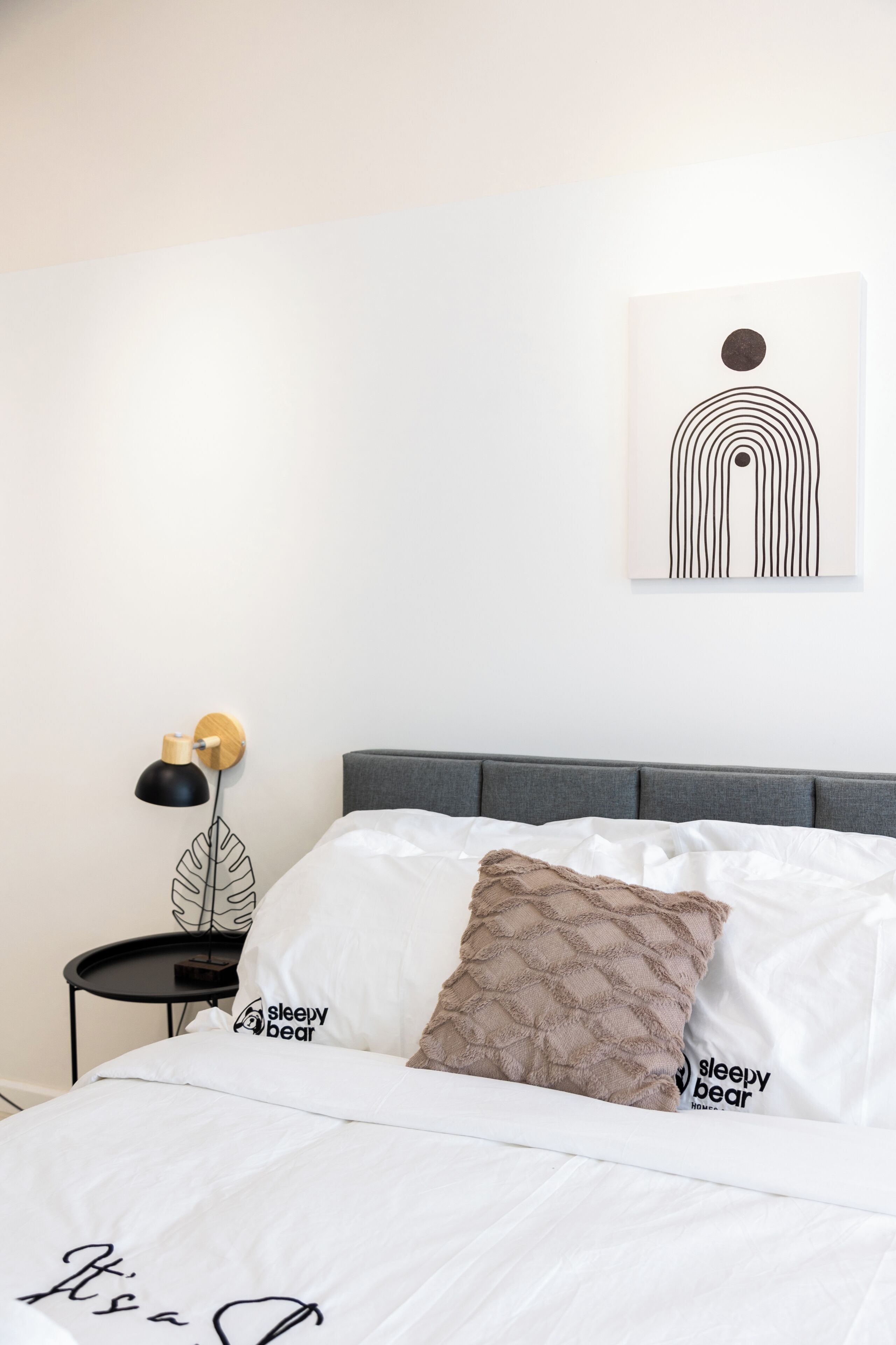 A neatly made bed with a pillow labeled 'sleepy bear,' a wall-mounted abstract art piece, and a bedside lamp on a small table.
