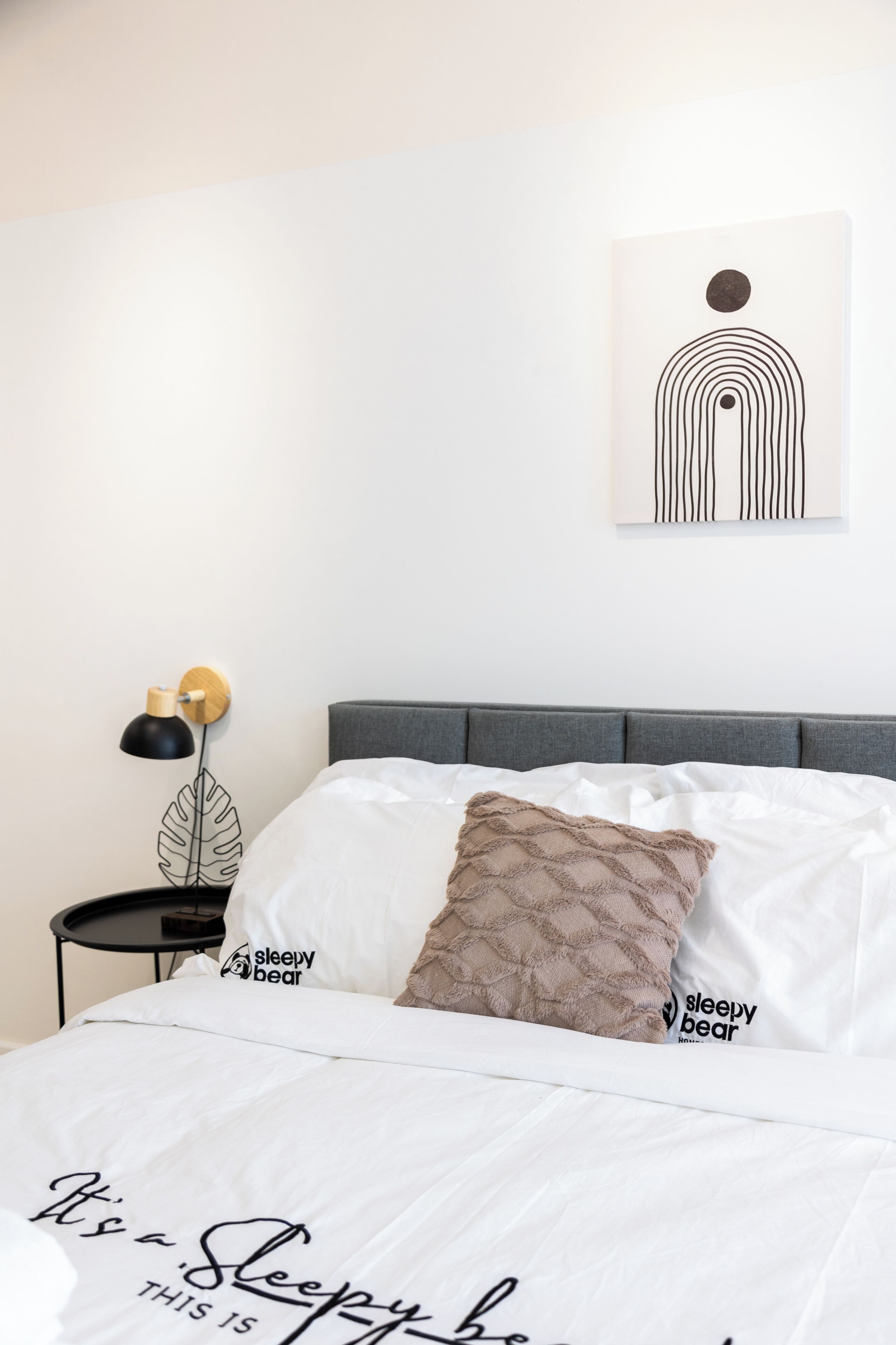 A neatly made bed with a white comforter and two pillows, a bedside table with a lamp, and a wall-mounted artwork.
