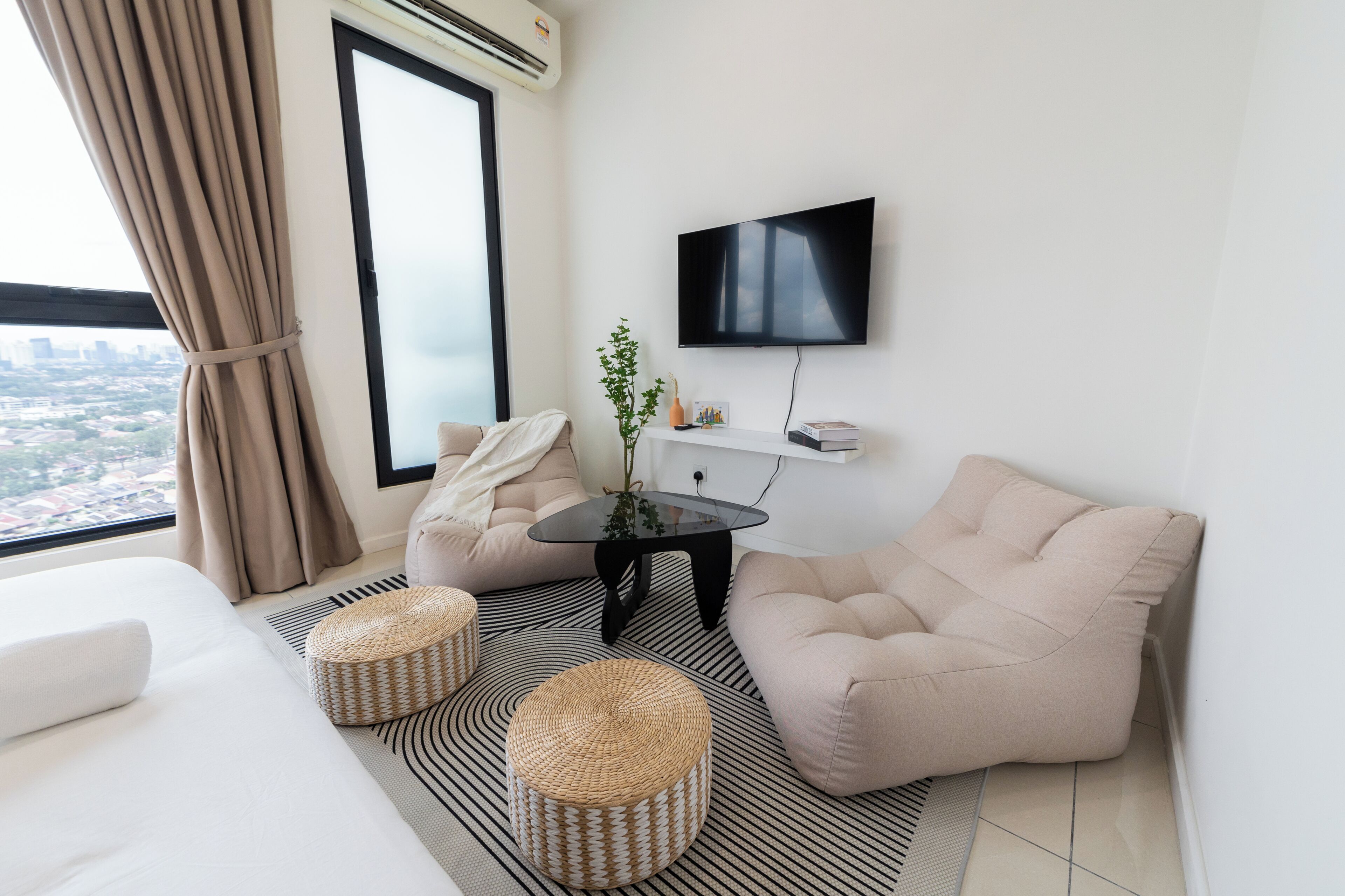 A modern hotel room with a large window, a flat-screen TV, a sofa, two ottomans, and a small table with a plant.