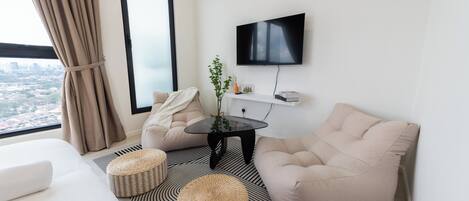 A modern hotel room with a large window, a flat-screen TV, a sofa, two ottomans, and a small table with a plant.