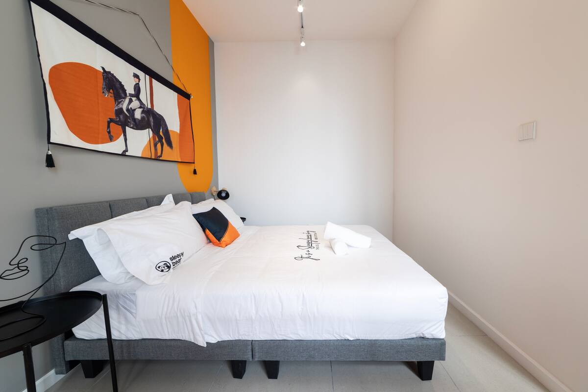 A neatly made bed with a white comforter and pillows, a gray headboard, a wall-mounted artwork, and a small bedside table with a lamp.