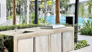 Una cocina al aire libre con pileta integrada y parrilla, rodeada de follaje y una piscina de fondo.