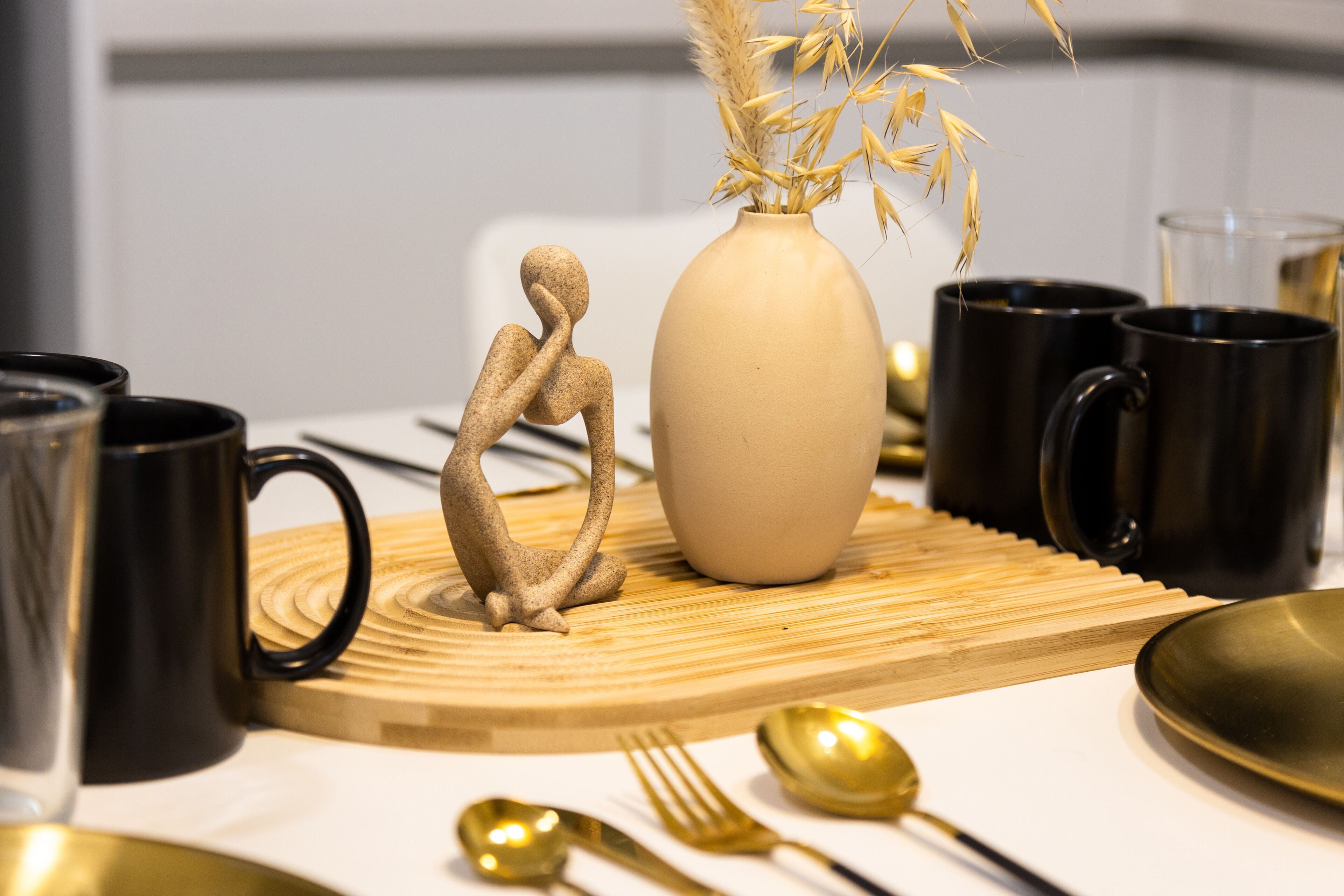 A table setting with a vase, black and gold mugs, a golden spoon, and a small sculpture.