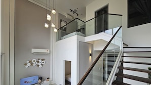 Una sala de estar moderna con un sofá, una mesa de centro y un televisor de pantalla plana. Se observa una escalera con barandillas de vidrio que conduce a un nivel superior.