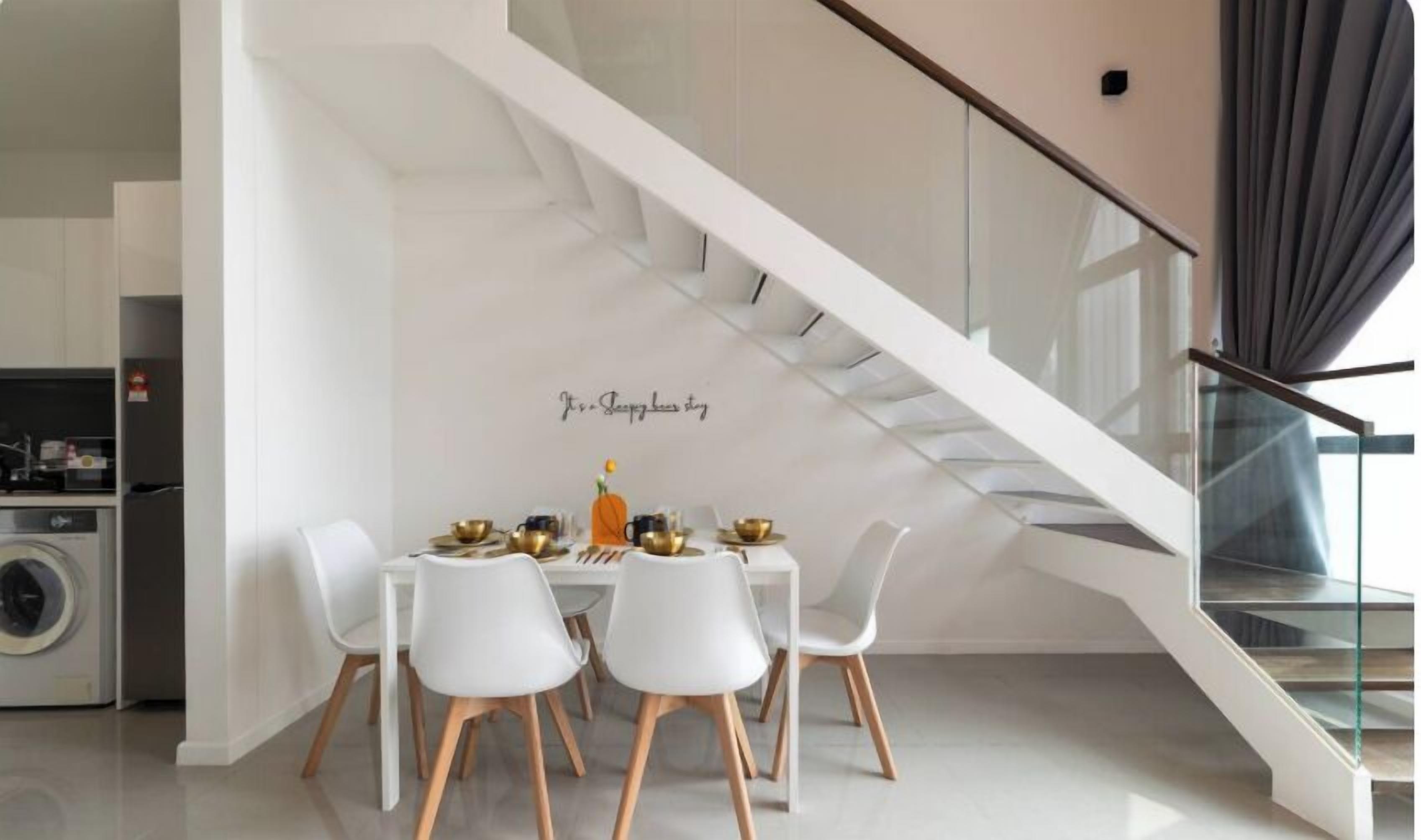 Una cocina moderna con zona de comedor, sillas blancas y una escalera con barandilla de vidrio.