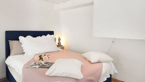 Una cama bien hecha con una manta rosa, almohadas blancas y una revista. Hay una pequeña estatua en una mesita de noche junto a un enchufe de pared.