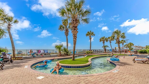 A pool area with lounge chairs, palm trees, and a clear sky. - Compass Cove Mariner 1415 (Myrtle Beach)