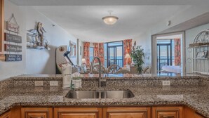 A kitchen with granite countertops, wooden cabinets, a stainless steel sink, and a view of the ocean through large windows. - Compass Cove Mariner 1415 (Myrtle Beach)