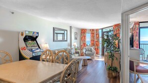 A living room with a wooden table, chairs, a sofa, a lamp, a plant, and a view of the ocean. - Compass Cove Mariner 1415 (Myrtle Beach)