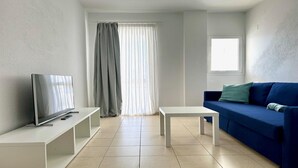A modern living room with a flat-screen TV on a white stand, a blue sofa with green cushions, a white coffee table, and a large window with sheer curtains. - refurbished holiday home very close to the beach (Solana Matorral)