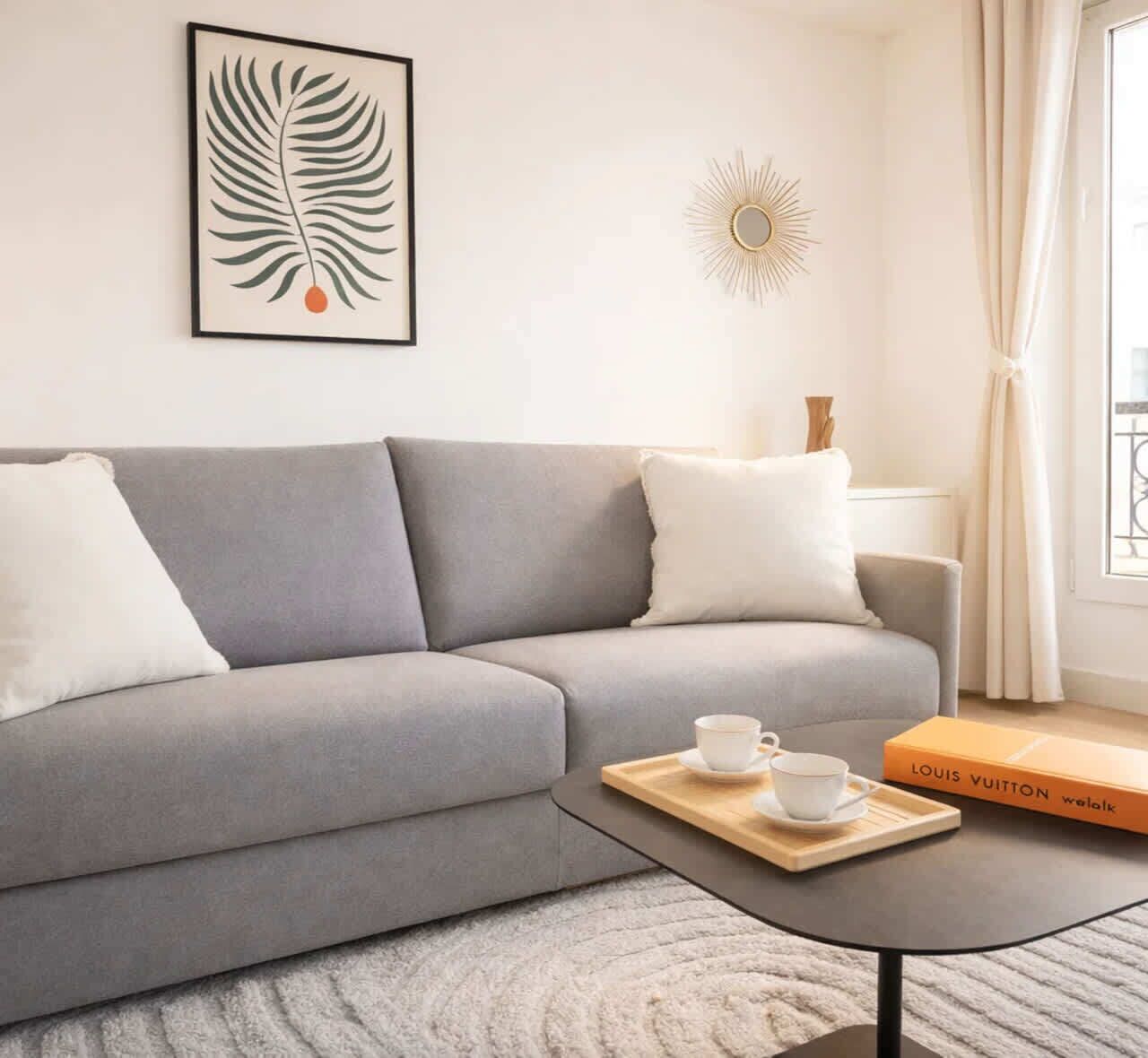 A modern living room with a grey sofa, a coffee table with a book, a framed leaf artwork, and a sunburst wall decoration.