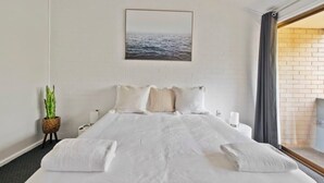 Une chambre d’hôtel avec un grand lit, du linge de lit blanc, une plante en pot, une table de chevet et une vue sur la mer par la fenêtre.