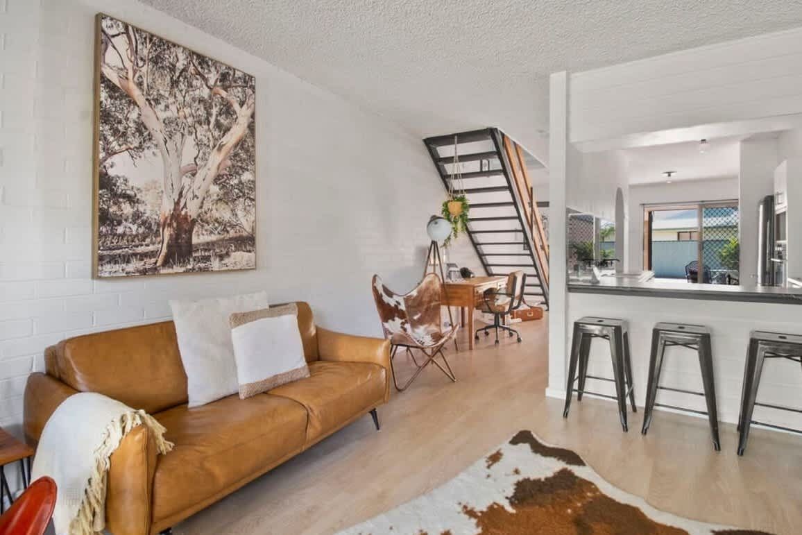 Un salon moderne avec un canapé en cuir marron, un coin repas avec une table et des chaises, une cuisine avec des tabourets de bar et un escalier.