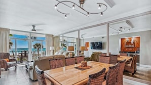 A spacious dining area with a large wooden table and wicker chairs, a kitchen with a TV, and a balcony with a view of the ocean. - 🏖️Gulf Front~Balcony~Pool~Hot Tub~Tennis Court~2BR Jade Gem🏖️ (Destin)