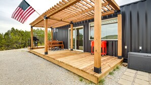 A covered wooden deck with a grill and outdoor seating, attached to a modern building with large windows and a black exterior. - Remote Strawn Container Home w/ Hot Tub! (Strawn)