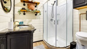 A bathroom with a glass-enclosed shower, a vanity with a mirror, and shelves with toiletries. - Remote Strawn Container Home w/ Hot Tub! (Strawn)