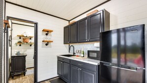 A compact kitchen with dark cabinetry, a black refrigerator, and a microwave. There are wooden shelves above the sink and a small countertop with a cutting board and bottles. - Remote Strawn Container Home w/ Hot Tub! (Strawn)