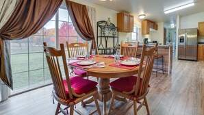 Un comedor con una mesa redonda de madera, seis sillas de madera con cojines rojos, una araña y una ventana con cortinas.