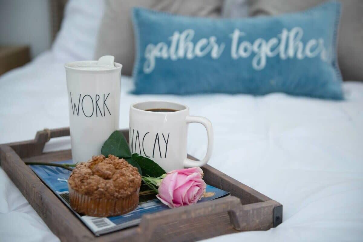 Una bandeja con un muffin, una taza de café con la etiqueta 'WORK' y otra taza de café con la etiqueta 'VACAY' sobre una cama con un almohadón que dice 'gather together'.