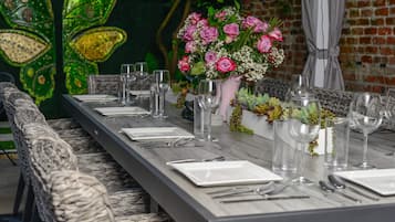 A long dining table set with plates, cutlery, and wine glasses, flanked by wicker chairs. A vase with pink flowers and greenery is placed on the table.