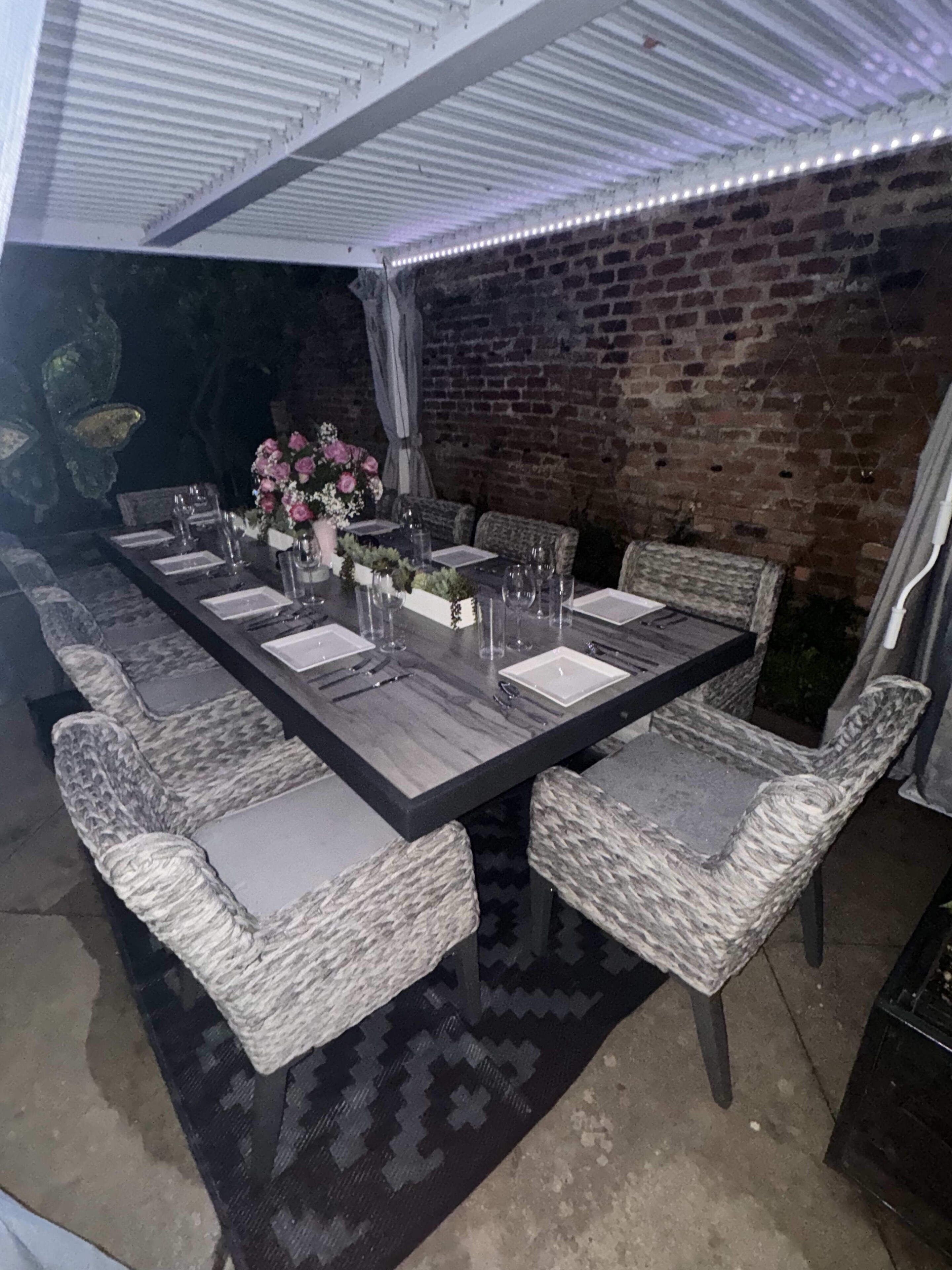 Un área de comedor al aire libre con un muro de ladrillo de fondo, una sección cubierta con techo blanco y una alfombra estampada.