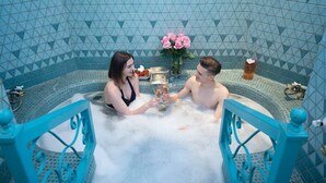 Ein Paar in einem türkisfarben gefliesten Whirlpool mit einem dekorativen Metallgeländer, einer Vase mit rosa Rosen und einer Flasche Orangensaft.