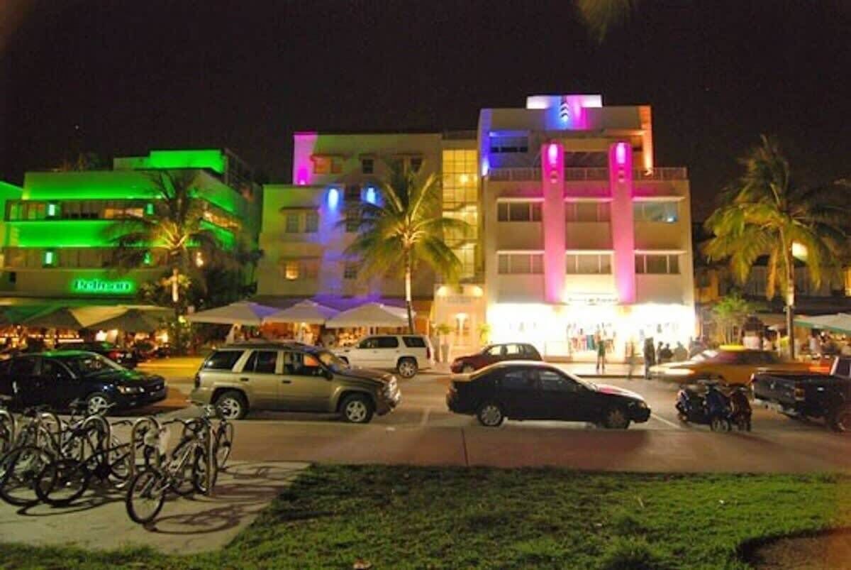 Un hotel de varios pisos con una fachada iluminada, un estacionamiento con coches y bicicletas, y palmeras en primer plano.