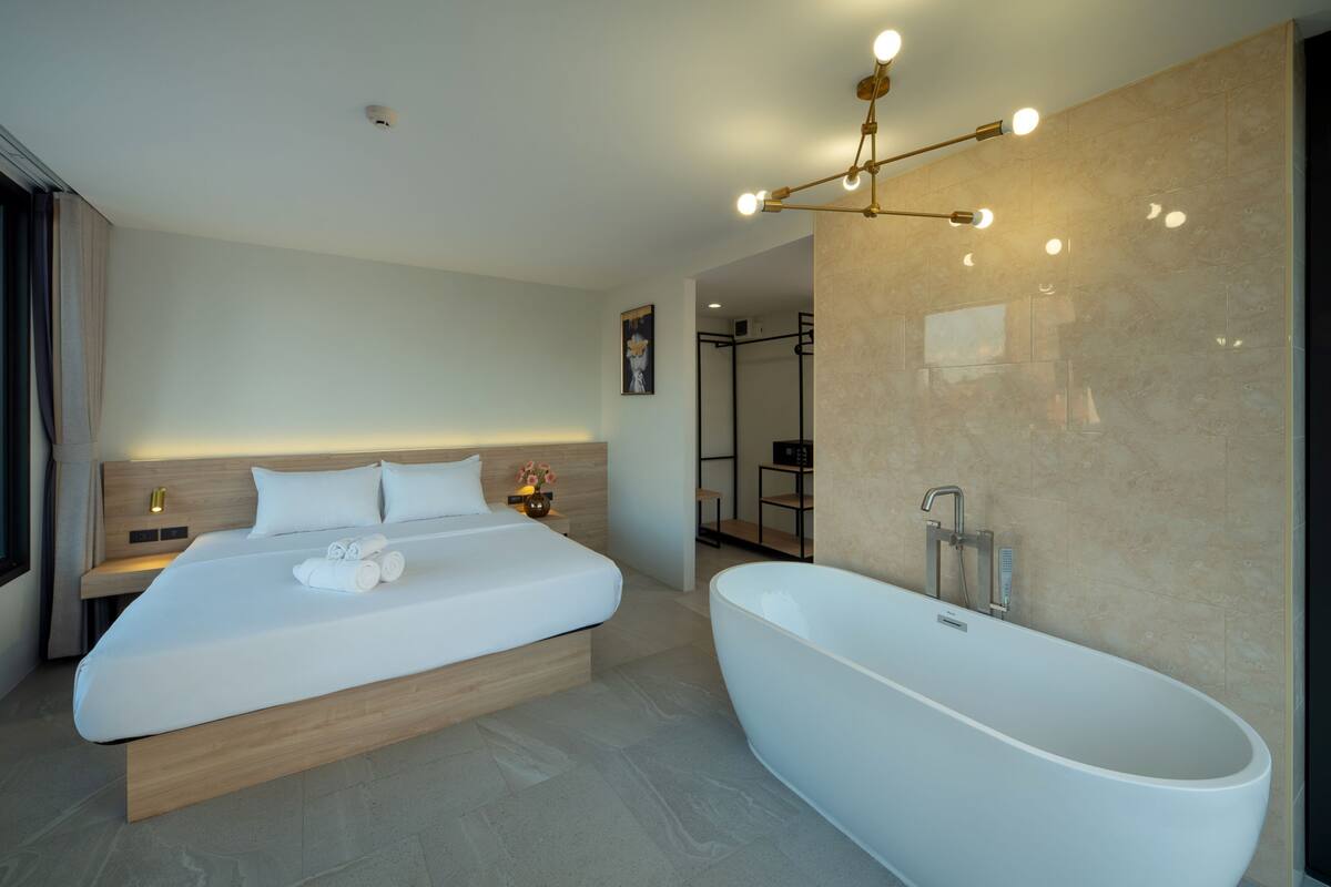 A modern bedroom with a large bed, a freestanding bathtub, and a bedside table with a lamp.