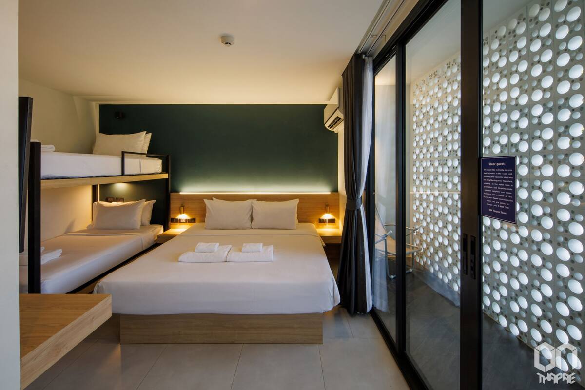 A hotel room with a large bed, two smaller beds, a wooden bedside table, a nightstand, and a glass partition with a decorative wall.