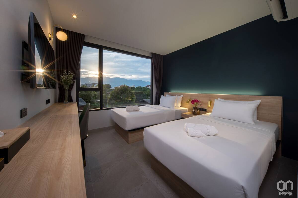 A modern hotel room with two beds, a desk, and a large window offering a city view.