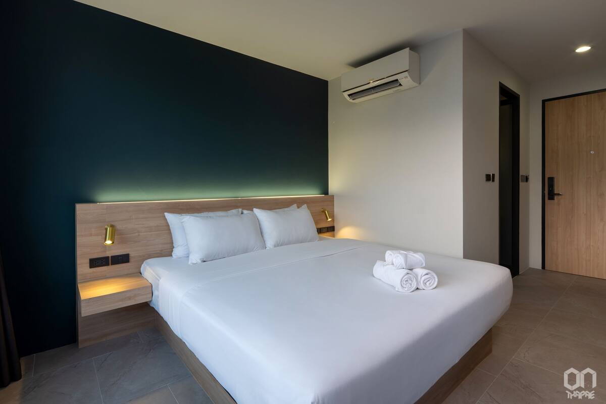 A modern hotel room with a large bed, a bedside table, an air conditioning unit, and a wooden door.