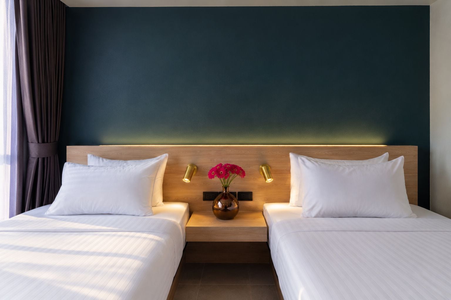 A hotel room with two beds, a nightstand with a vase of flowers, and a wall-mounted light fixture.
