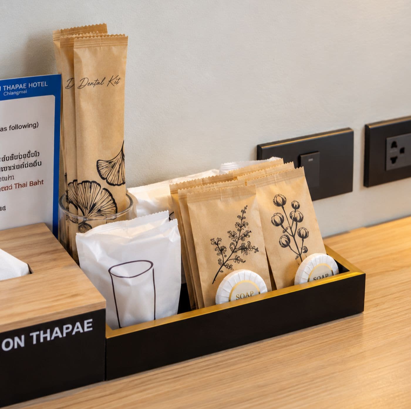 Un espace d’accueil d’hôtel avec un comptoir en bois, une enseigne indiquant « Thapae Hotel », et une boîte contenant des articles de toilette, notamment du savon et des serviettes en papier.