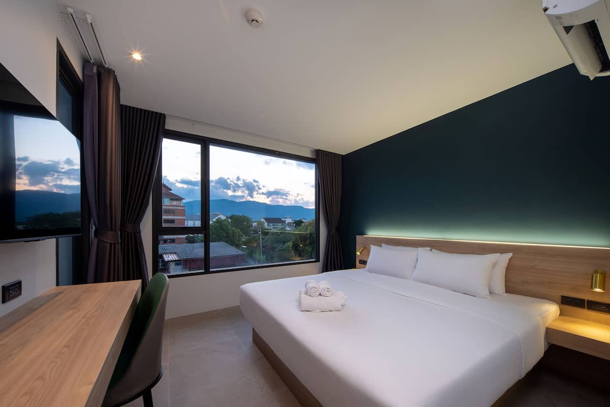 A hotel room with a bed, a desk, a chair, a TV, and a window with a view of buildings and mountains.
