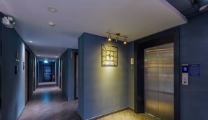 Un couloir d’hôtel moderne, avec une grande horloge murale, un ascenseur et un passage menant à une porte.
