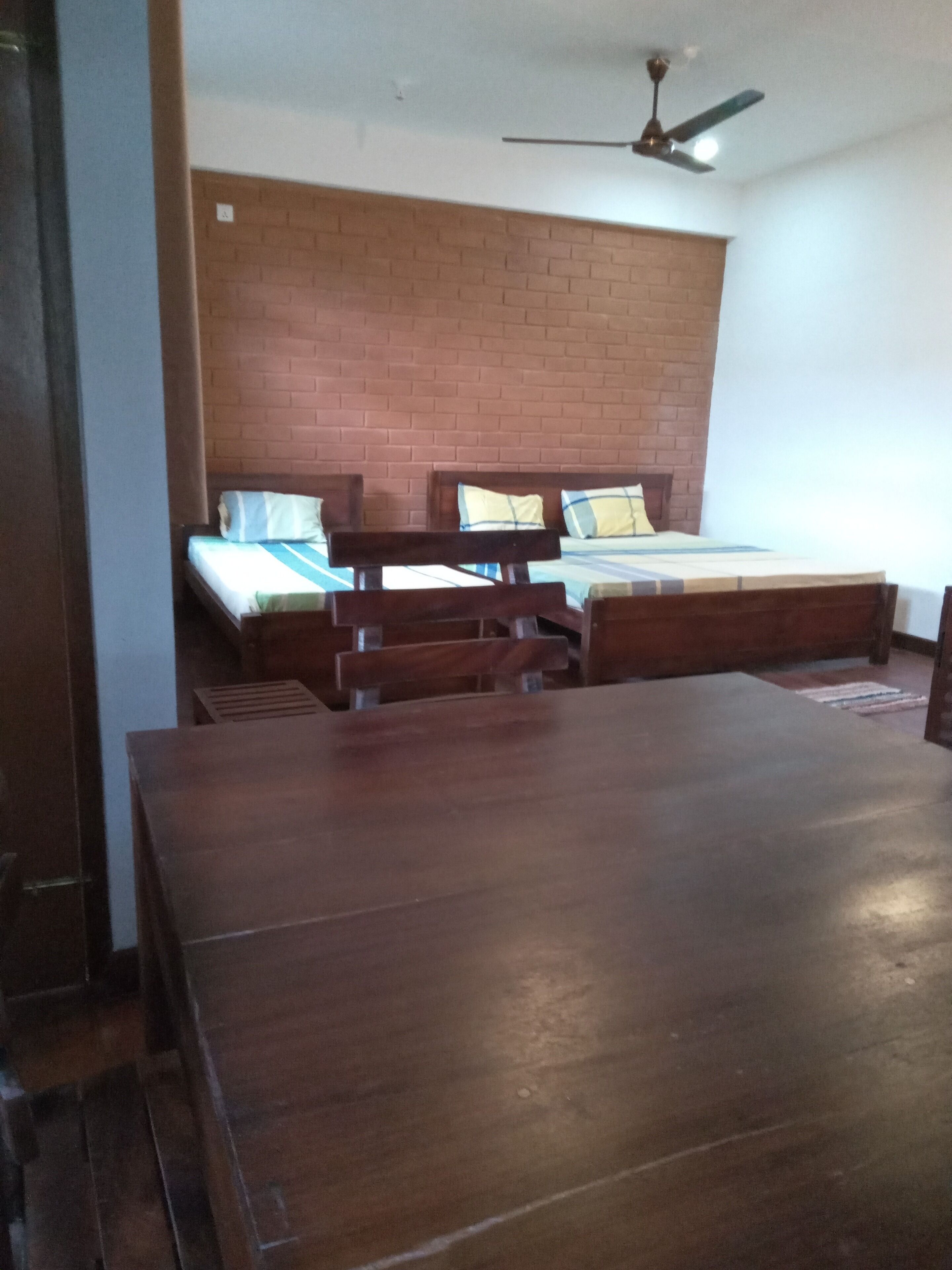 A room with a large wooden table, two beds with wooden frames, a brick wall, and a ceiling fan.