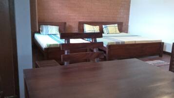 A room with a large wooden table, two beds with wooden frames, a brick wall, and a ceiling fan.